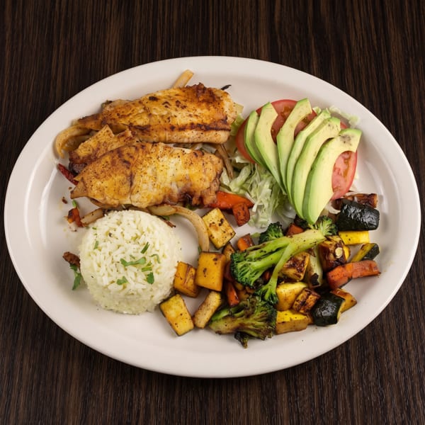 Plate of grilled fish with rice, roasted vegetables, avocado, tomato, and lettuce salad on a dark wooden table