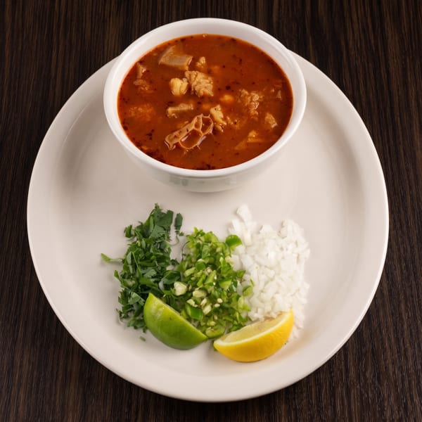 Bowl of red soup with meat, served with cilantro, chopped onions, green chilies, lime, and lemon wedges on a white plate.