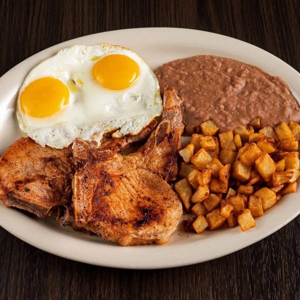 Plate of grilled pork, two sunny-side-up eggs, refried beans, and golden diced potatoes on a dark wooden table.